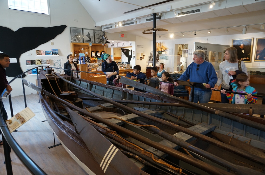 A crowd of adults and children surround a historic whaleboat with a museum educator in the museum gallery.