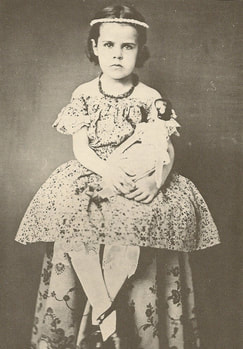 Uncolored photo of young girl about 5 years old sitting with dress and necklace, legs crossed at the ankle, and somewhat serious staring face, holding a doll
