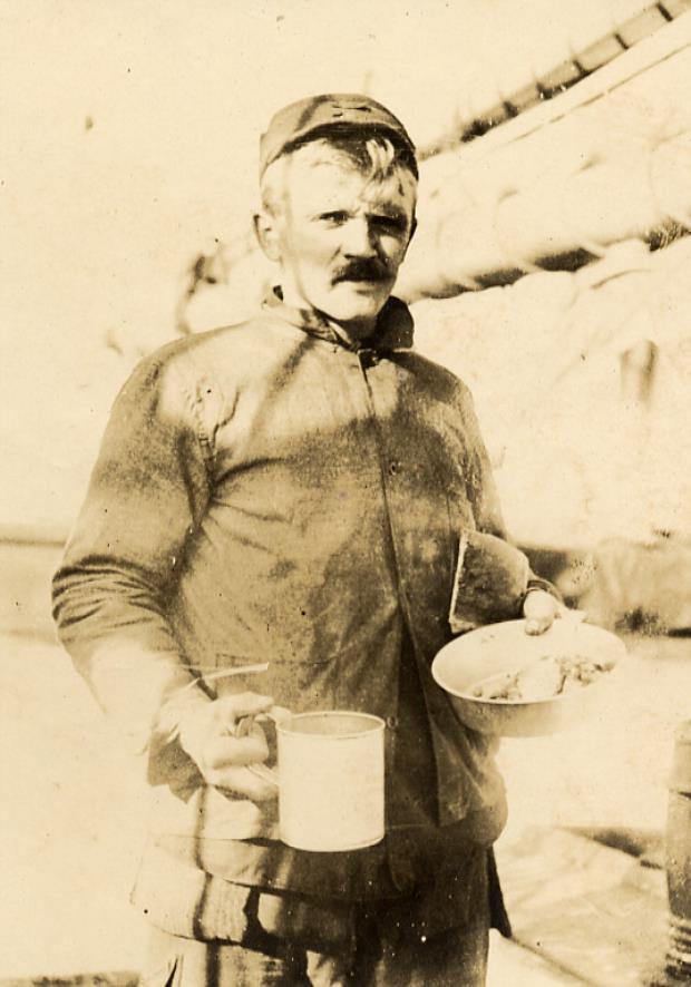 A whaler faces the viewer holding a tin cup and plate filled with food. 