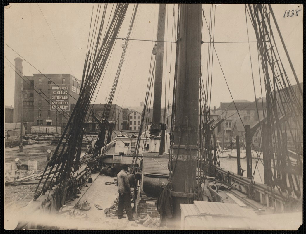Black and white photo of man on deck of whaling ship docked in New Bedford, Massachusetts; man stands by the ship's tryworks; buildings in background, Humphrey's Cold Storage and Old Colony Creamery building at left.e
