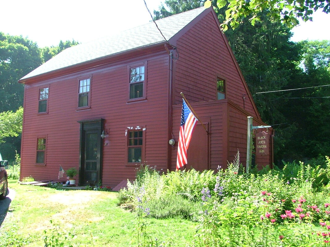 A sunny photo of a colonial home with red wooden siding surrounded by a garden and american flag, with a sign that says 