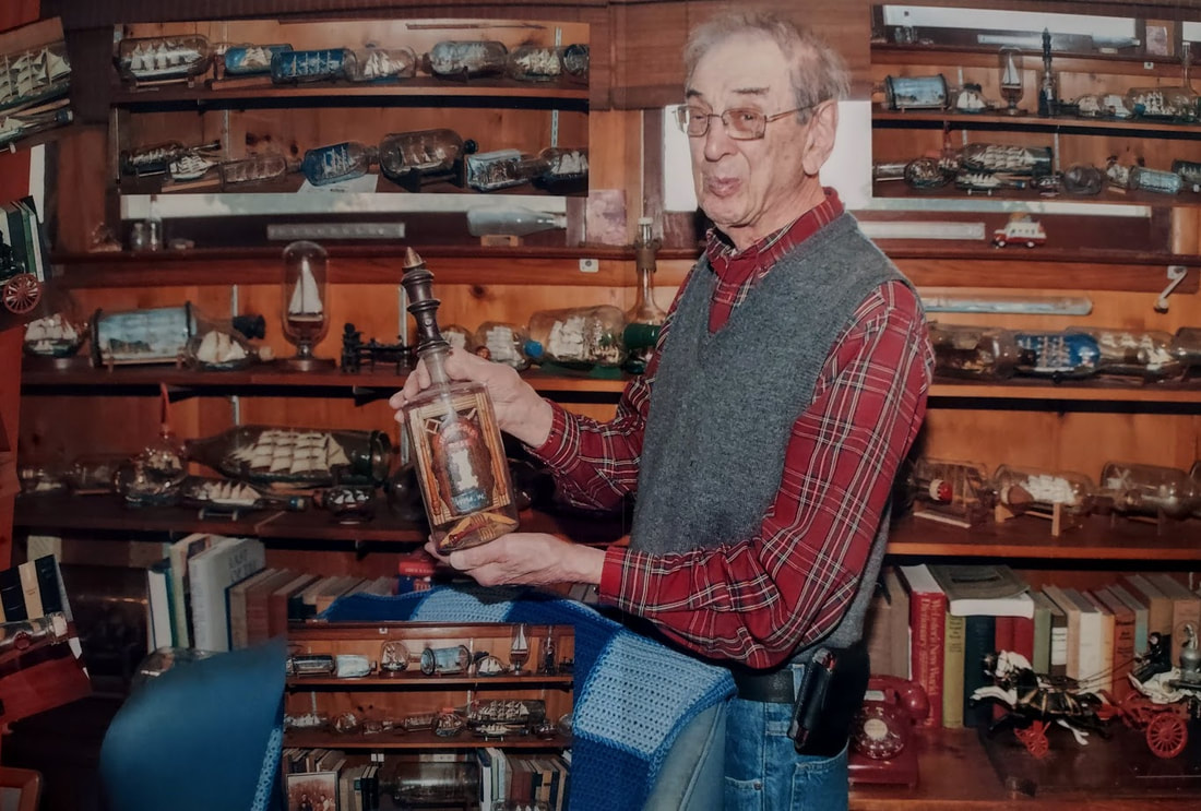 A standing senior man wearing a vest holds a bottle in front of his ship in a bottle collection