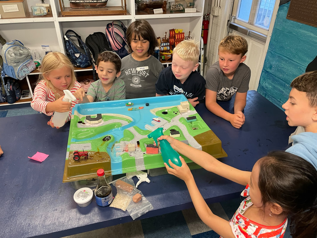 Children crowd around a table with a learning model showing the flow of water in a town setting and into a bay. Children hold spray bottles