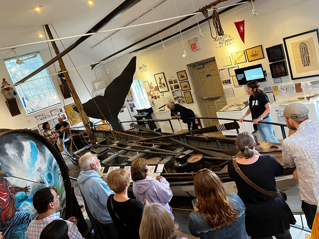 A museum educator stands with a crowd around the museum's historic wooden whaleboat as she leads a discussion