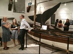 Adults leaning around a displayed wooden brown whaleboat in the museum gallery.