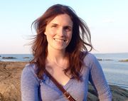 Smiling Brenna with beach background. Her long, reddish hair frames her fair skin.