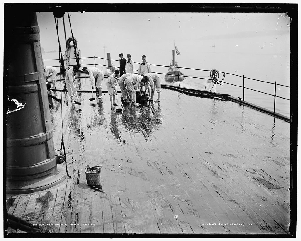 Black and white photo of 6 men bending over a bucket to scrub a wet deck.